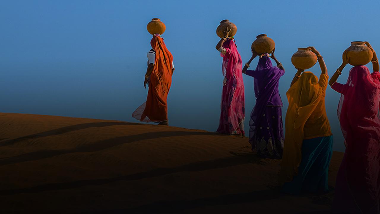 Women carrying pots of water across a desert.