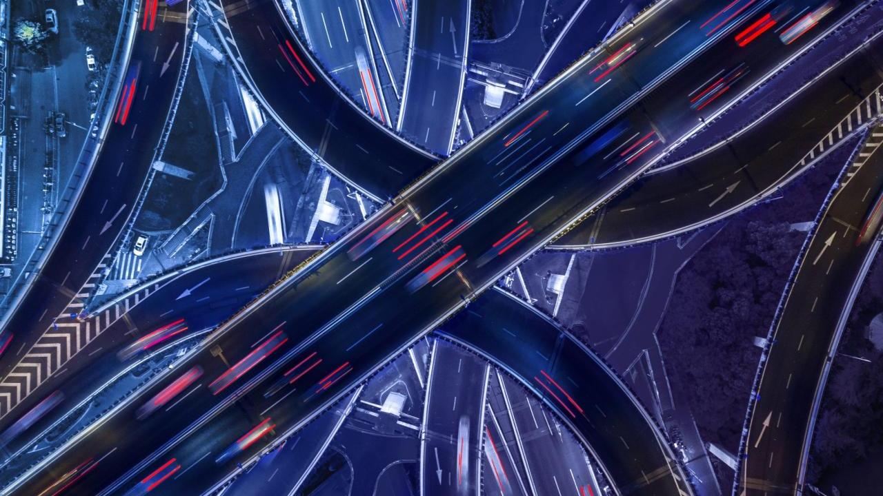 Nighttime aerial view looking down at a busy, intersecting highway with many ramps. Blurred vehicles on the roads.