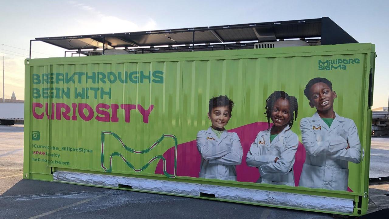A green shipping container turned mobile science lab features three young scientists and text reading, "Breakthroughs Begin With Curiosity."