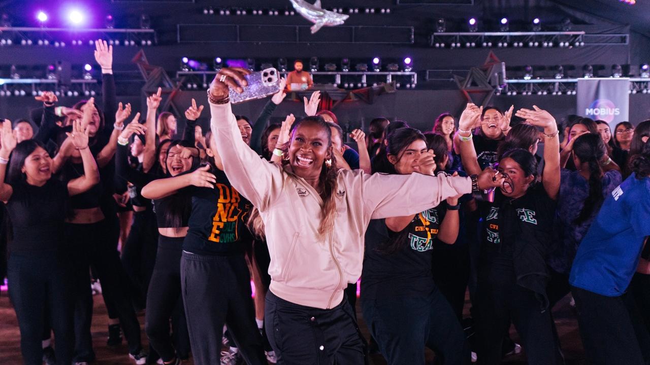 A person in a light jacket holds up a phone for a selfie while surrounded by a large group dancing with raised arms inside a concert-style venue with stage lighting overhead.