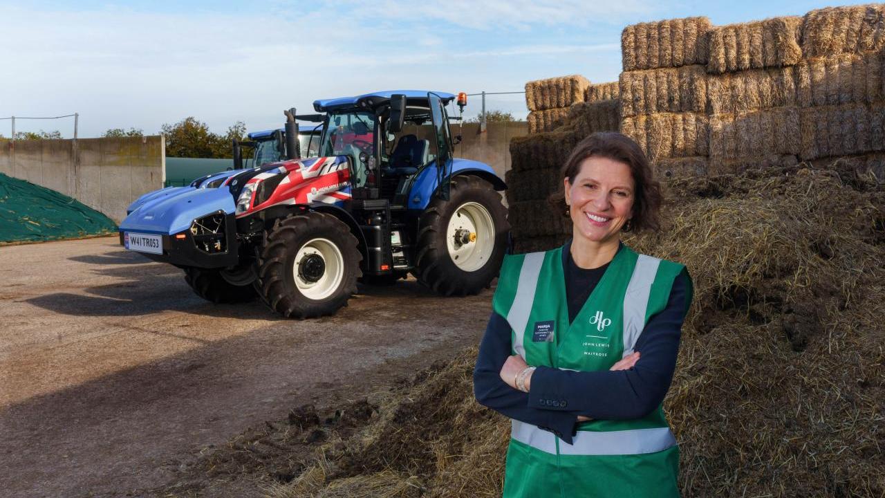 Person smiling with their arms folded, stood in front of a tractor and hay bales 
