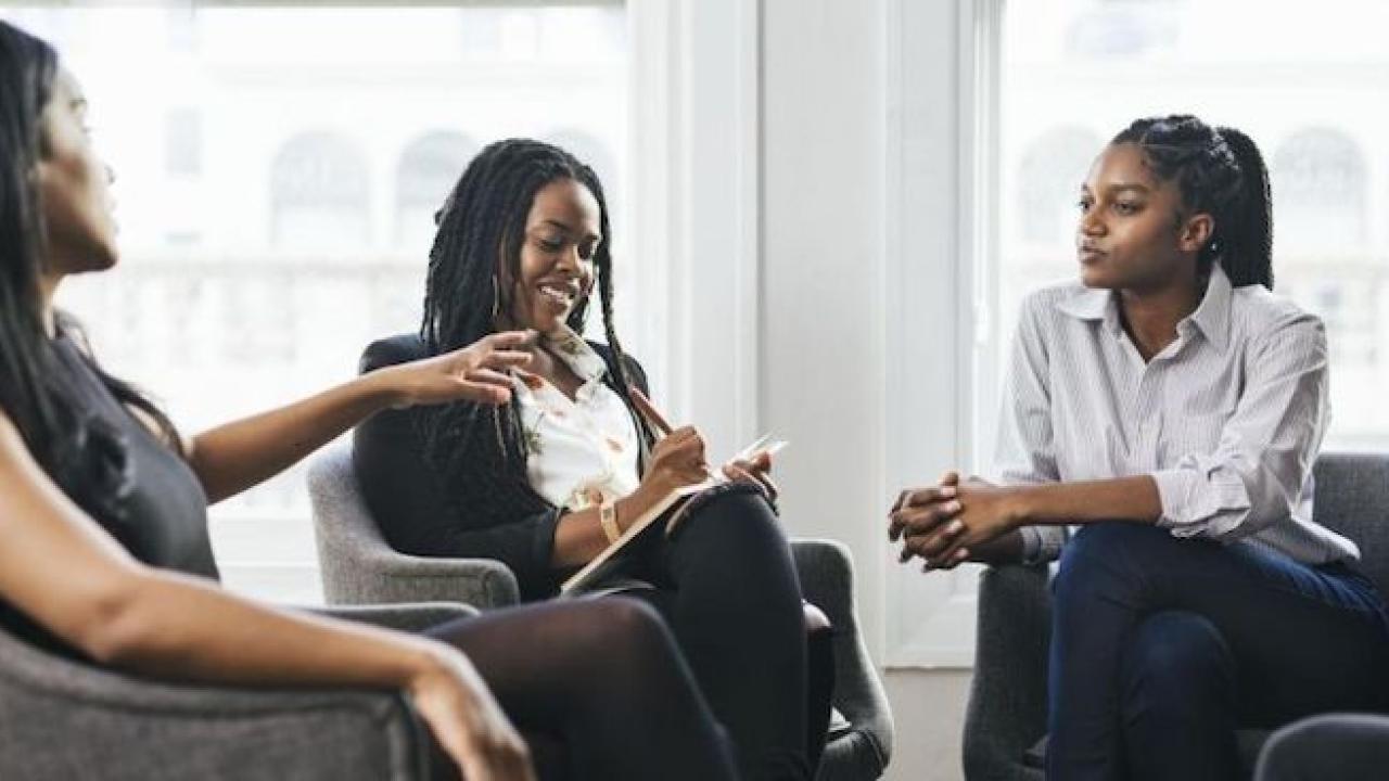 Three African American women in business attire are seated in a semi-circle having a discussion. The woman in the middle is taking notes on a pad. 