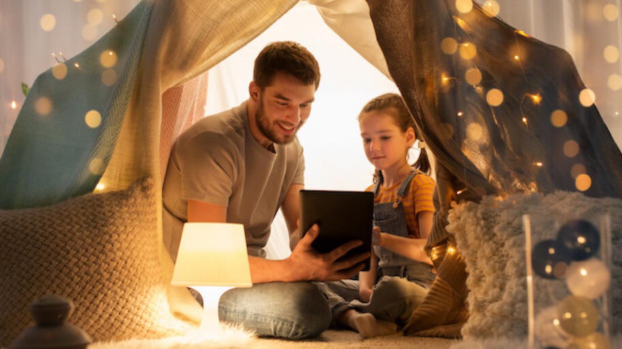 Father and daughter seated behind a curtain and looking at the screen of a laptop.