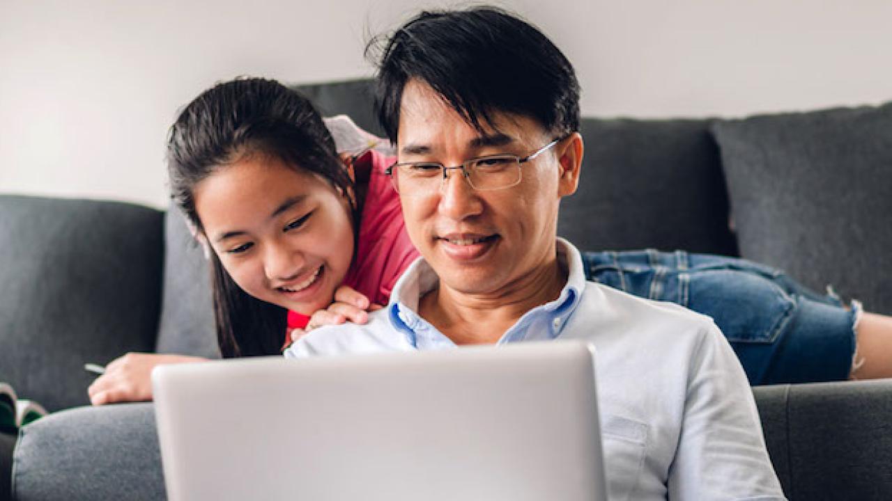 Father and daughter (daughter is sitting on a couch and dad is sitting on the floor) are looking at a laptop screen 