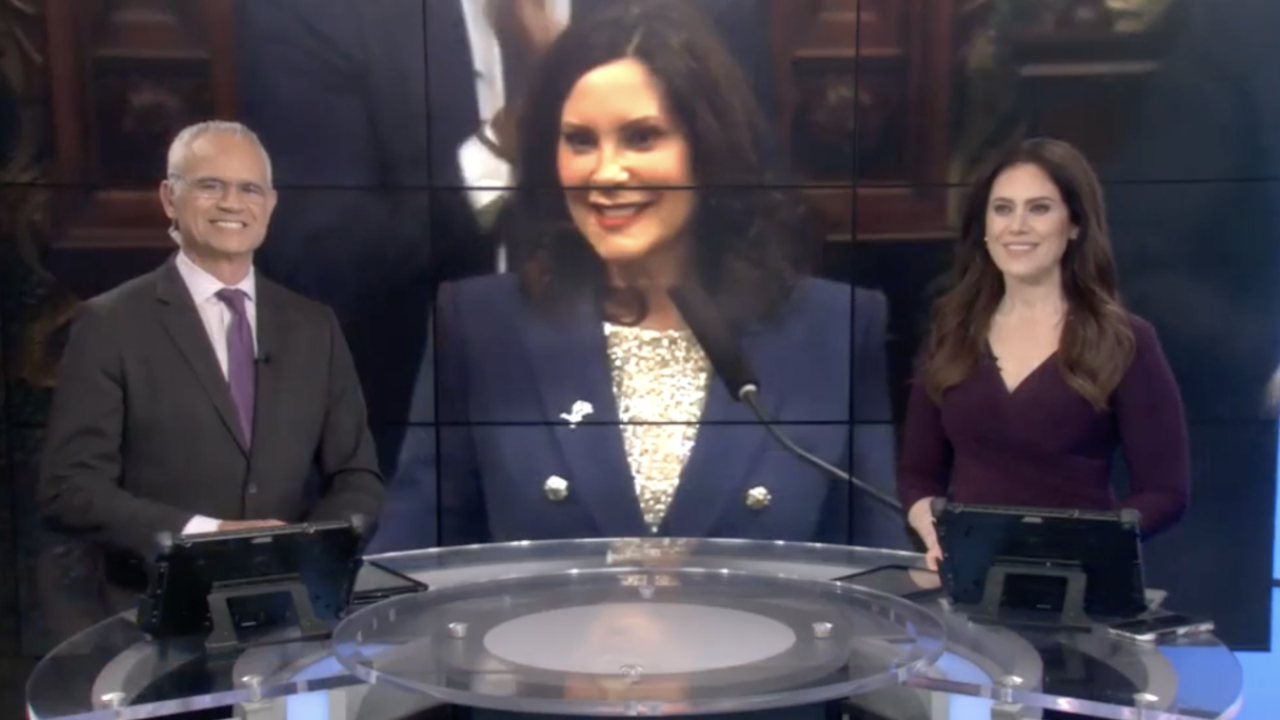 Two news hosts sat at as desk with a screen behind them