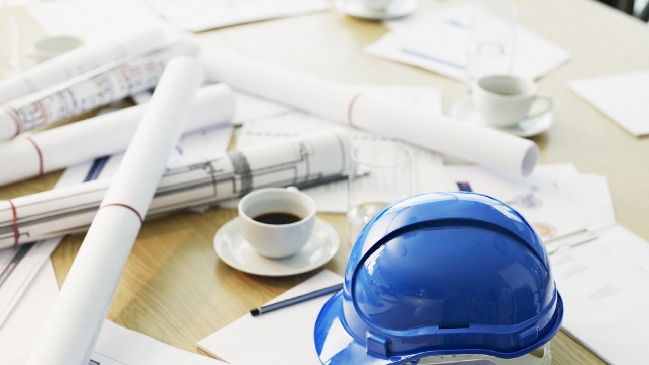 Desk littered with rolls of paper, a coffee cup, and a hard hat