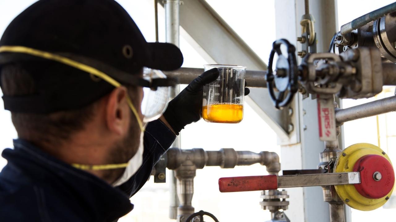 a person wearing protective glasses, mask and gloves holding a beaker of clear yellow liquid. Industrial pipes and valves in front of them.