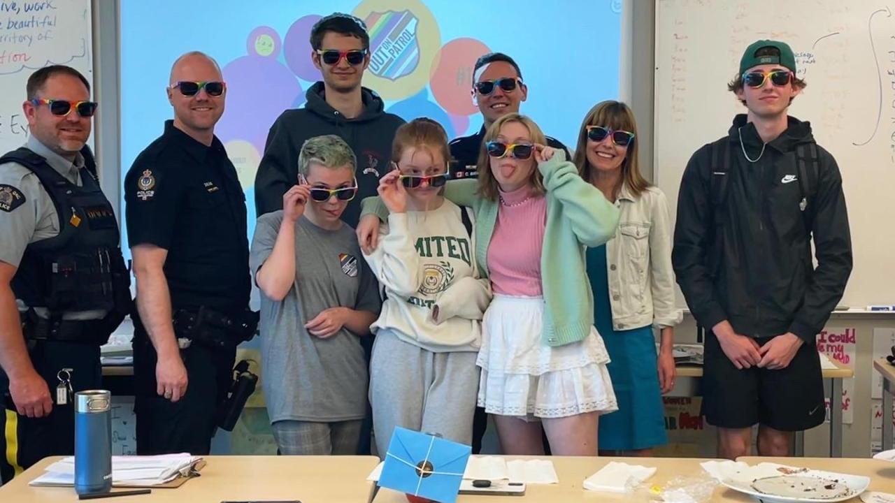 A group of adults and kids all wearing rainbow-rimmed glasses in a classroom setting. Some adults in police uniform.