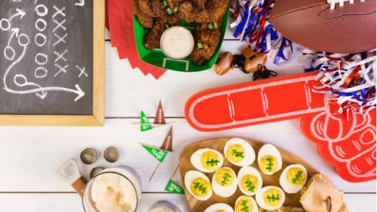 Party materials on a table for Super Bowl Sunday: Football, hard boiled eggs decorated like footballs, wings and a small chalk board with a play written in chalk.