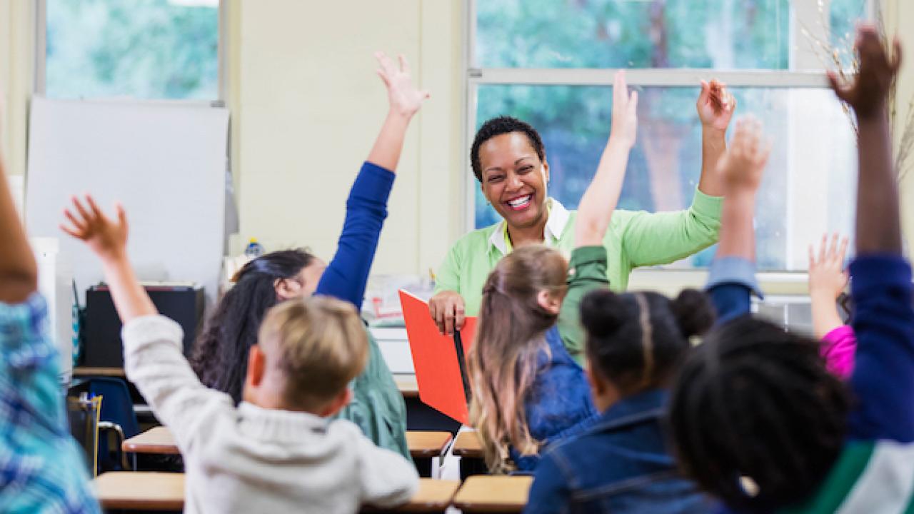 Female teacher in front of her classroom of children. The children have their hands raised.