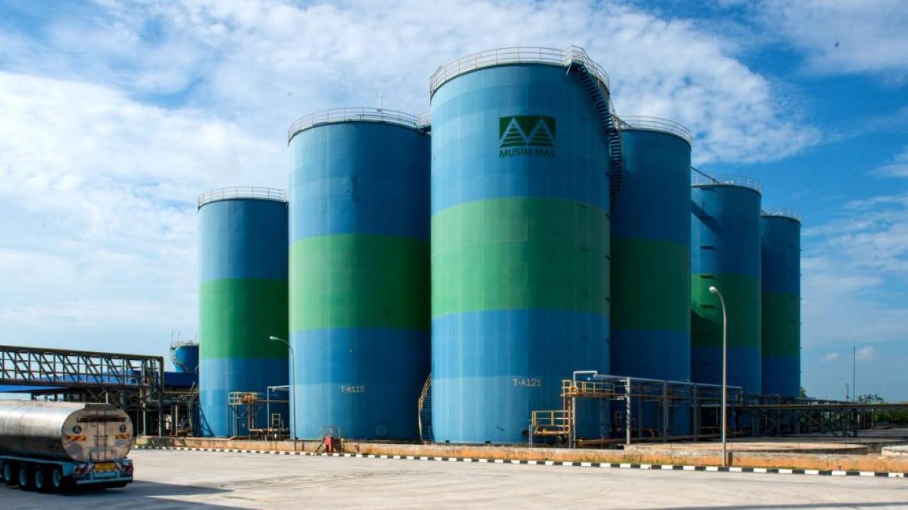 Towering holding tanks with Musim Mas logo. A semi truck driving by them. A blue cloudy sky above.