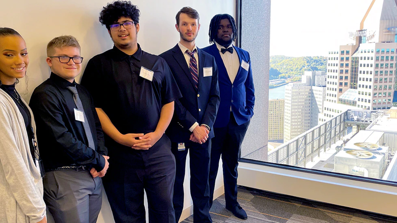 Five students in business style clothing standing in a hallway with a large window at the end overlooking a city landscape.