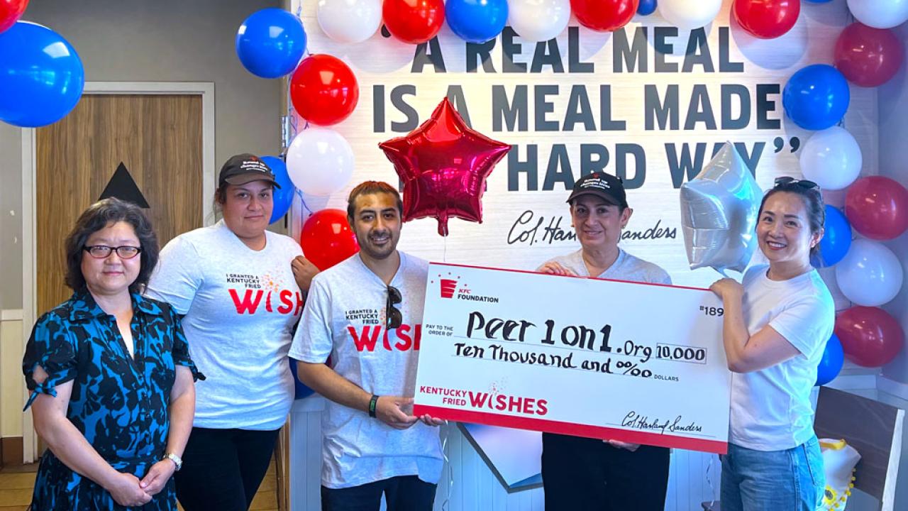 Group of people stood in front of a wall with red, white and blue balloons holding a Kentucky Fried Wishes funding cheque