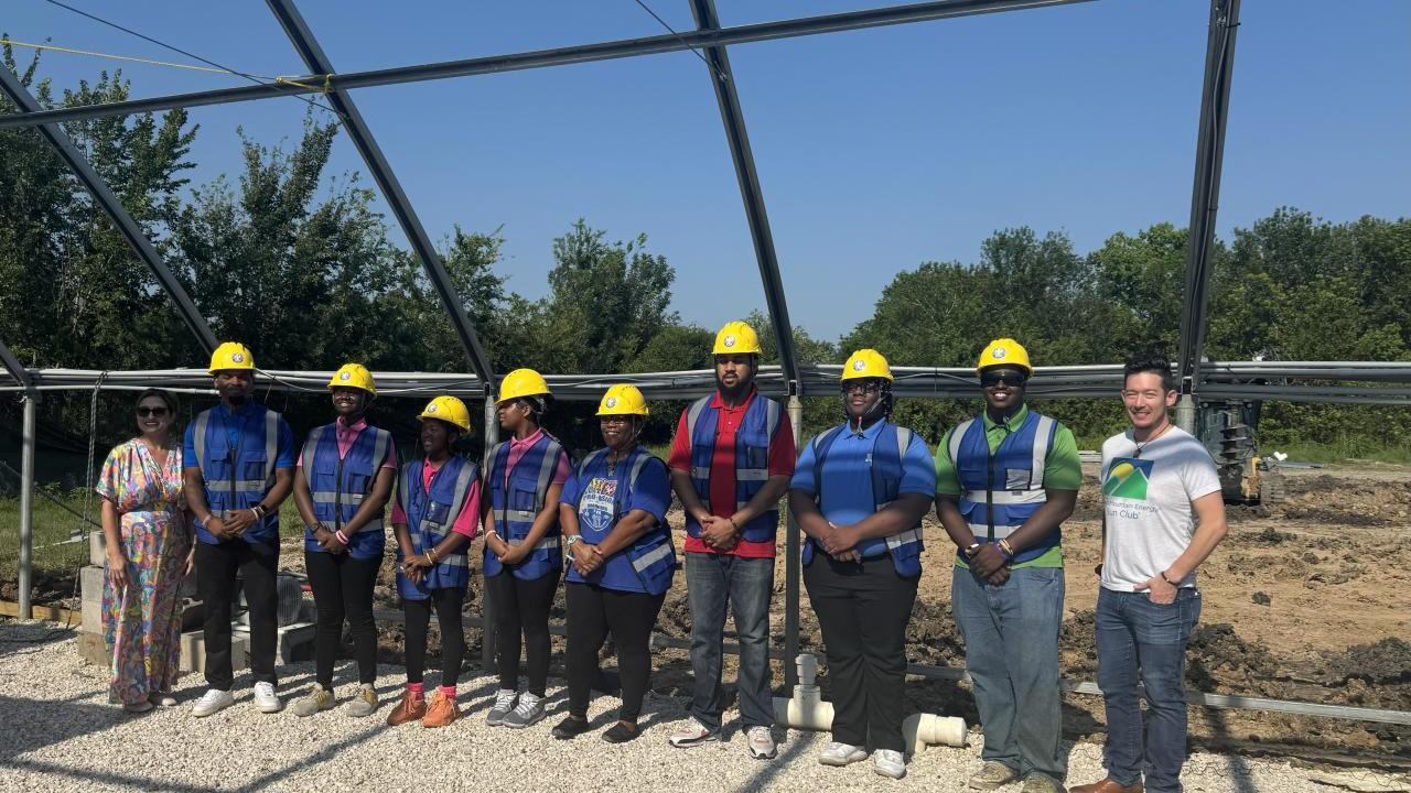 Group of people in greenhouse structure, wearing blue construction vests and yellow hard hats