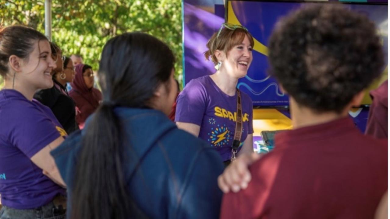 A woman in a MilliporeSigma Spark T-Shirt smiles alongside students. 