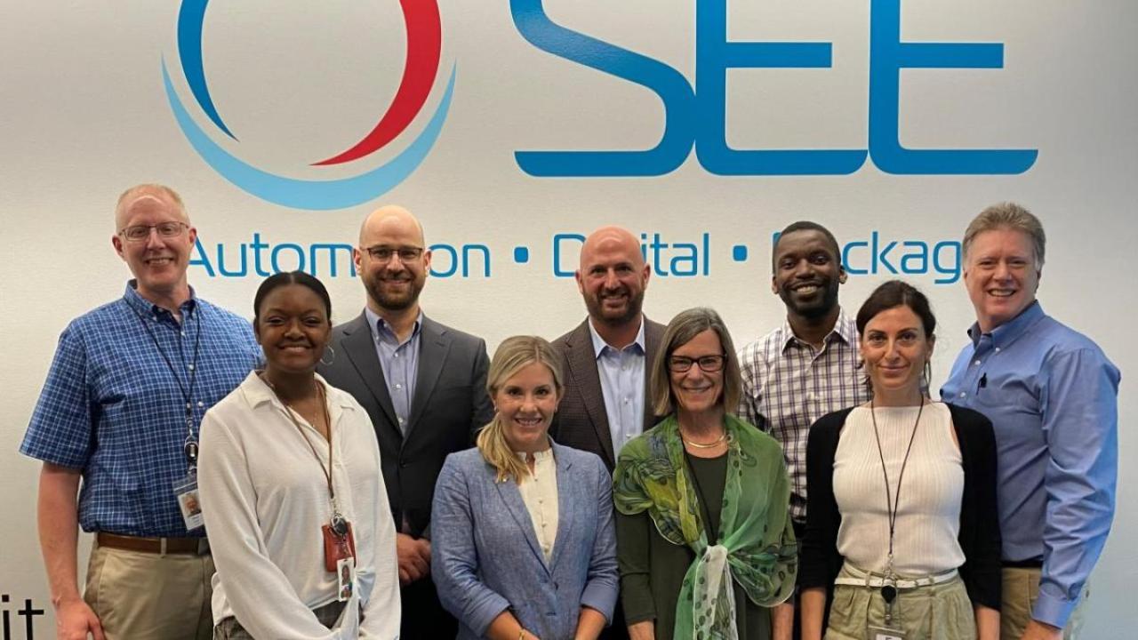 A group of people stand in front of the SEE company logo facing the camera.