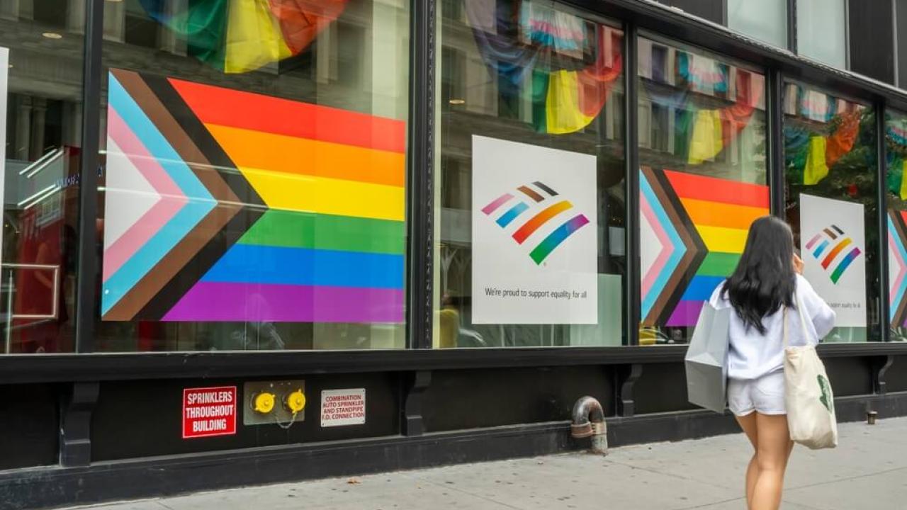 Pride flags shown in the windows of business on a city street. A woman is walking down the street with a shopping bag.
