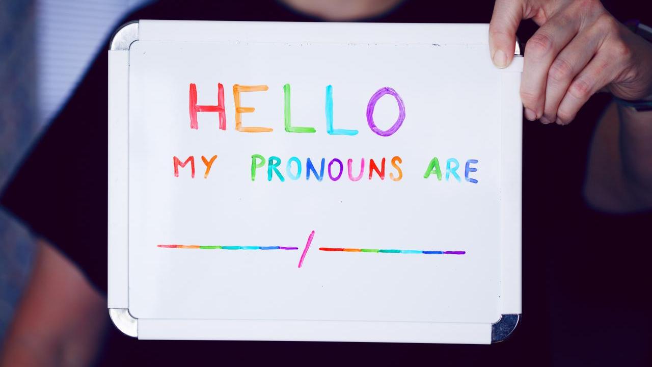 A person holding a sign with rainbow letters "Hello my pronouns are _/_"