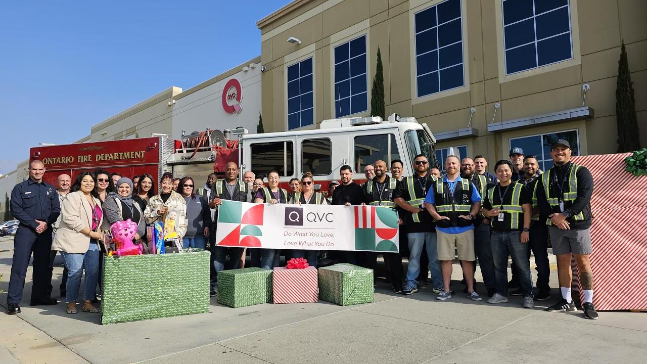 A mix of team members from Qurate Retail Group are standing outside a QVC facility in front of a firetruck along with fire fighters. A few people are holding up a banner which reads "QVC Do What You Love, Love What You Do". They are also surrounded by wrapped gift boxes and holding children's toys as part of a holiday toy drive event.  