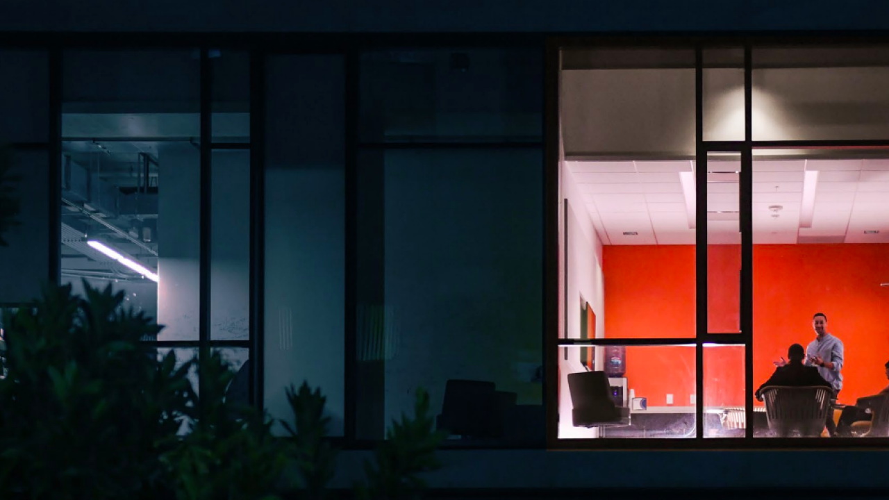 Looking in from the outside at two people talking in a lit office space, other spaces in the building are dark.