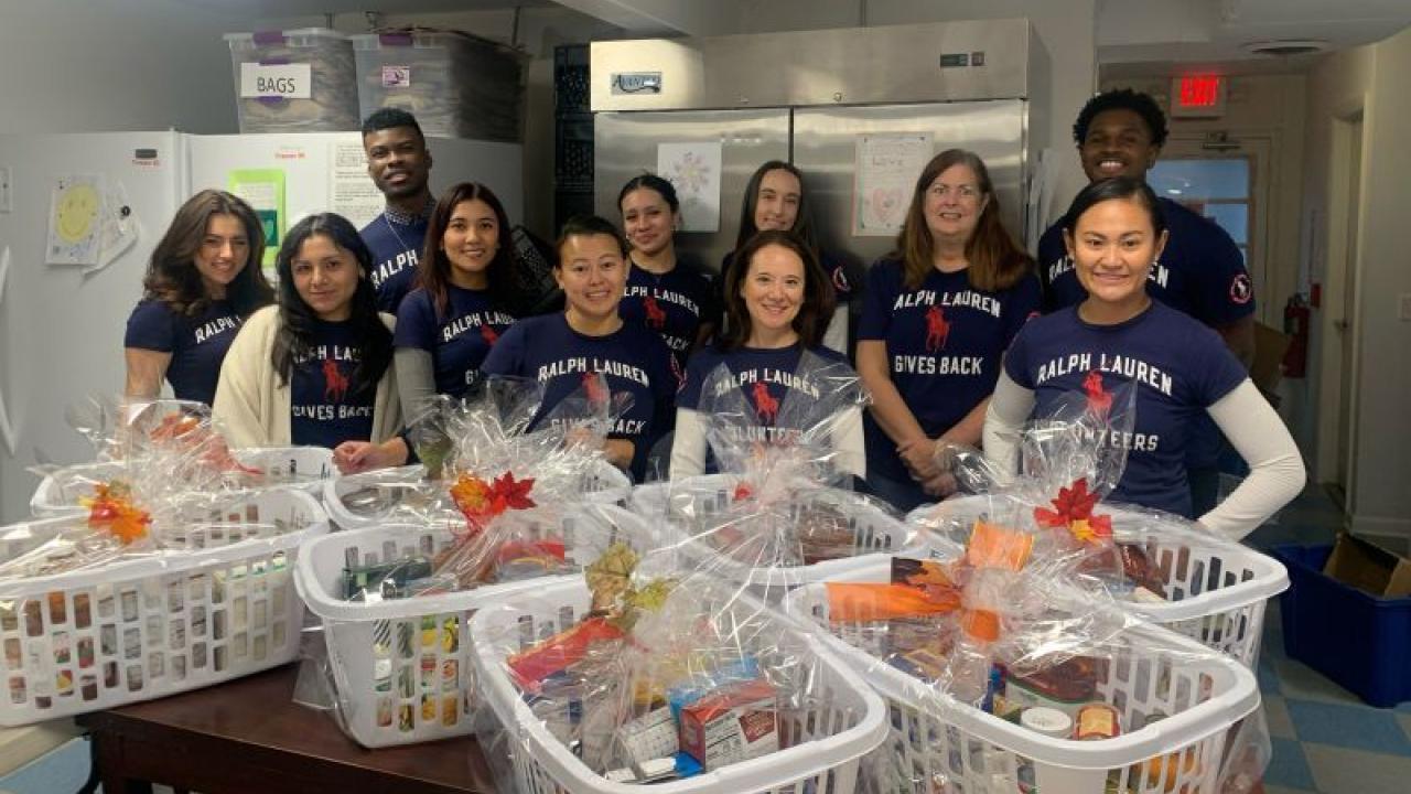 A group of people standing in a kitchen, large baskets of food items in front of them. All wearing "Ralph Lauren Volunteer" t-shirts.
