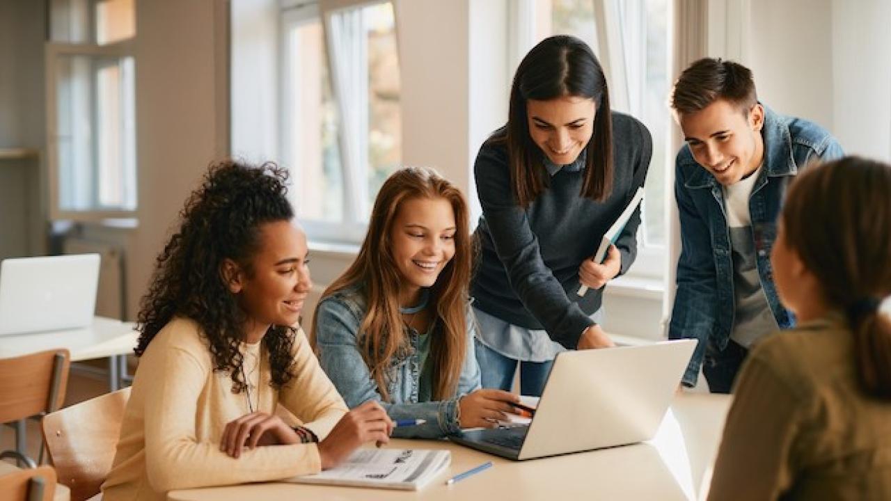 Teacher working with a group of four students who are seated behind a laptop.
