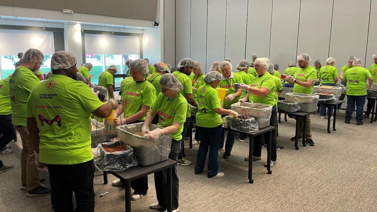 Volunteers wearing bright green SPARK™ t-shirts, hairnets, and gloves assemble meal kits.