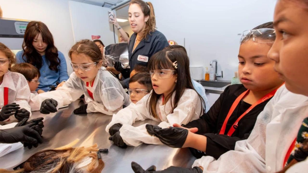 Children and adults in protective lab gear surround a table.