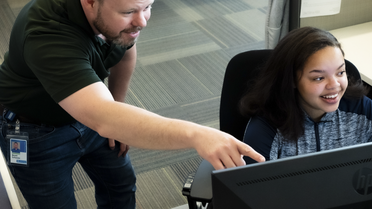 Person sat at a desk in front of a computer screen with someone stood next to them pointing at the monitor
