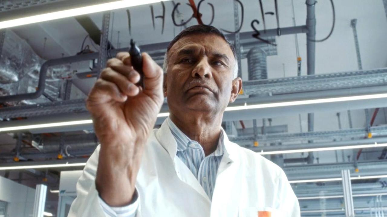 Dr. Ponnusamy wears a lab coat writing out chemical compounds on glass with a marker.