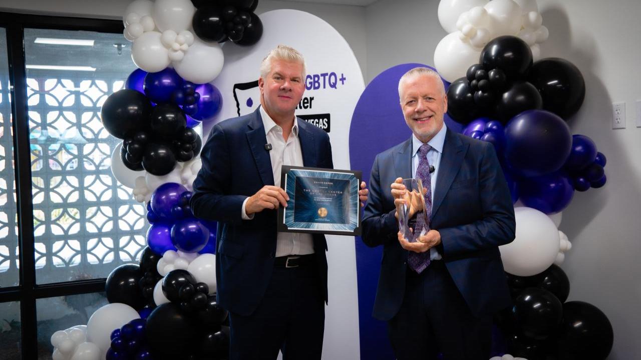 Two men standing in front of balloons. One is holding a framed certificate and one is holding an award.