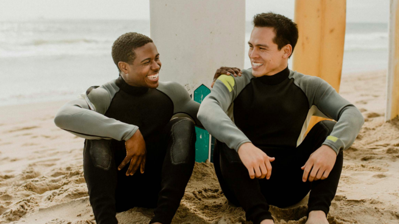 two men sitting on the beach wearing wet suites for surfing