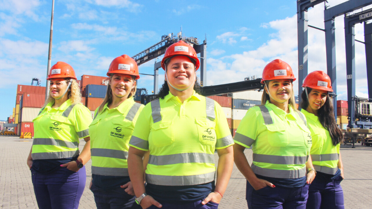 Five women wearing safety gear stand in front of container cranes and stacked shipping containers at DP World’s Santos terminal in Brazil.