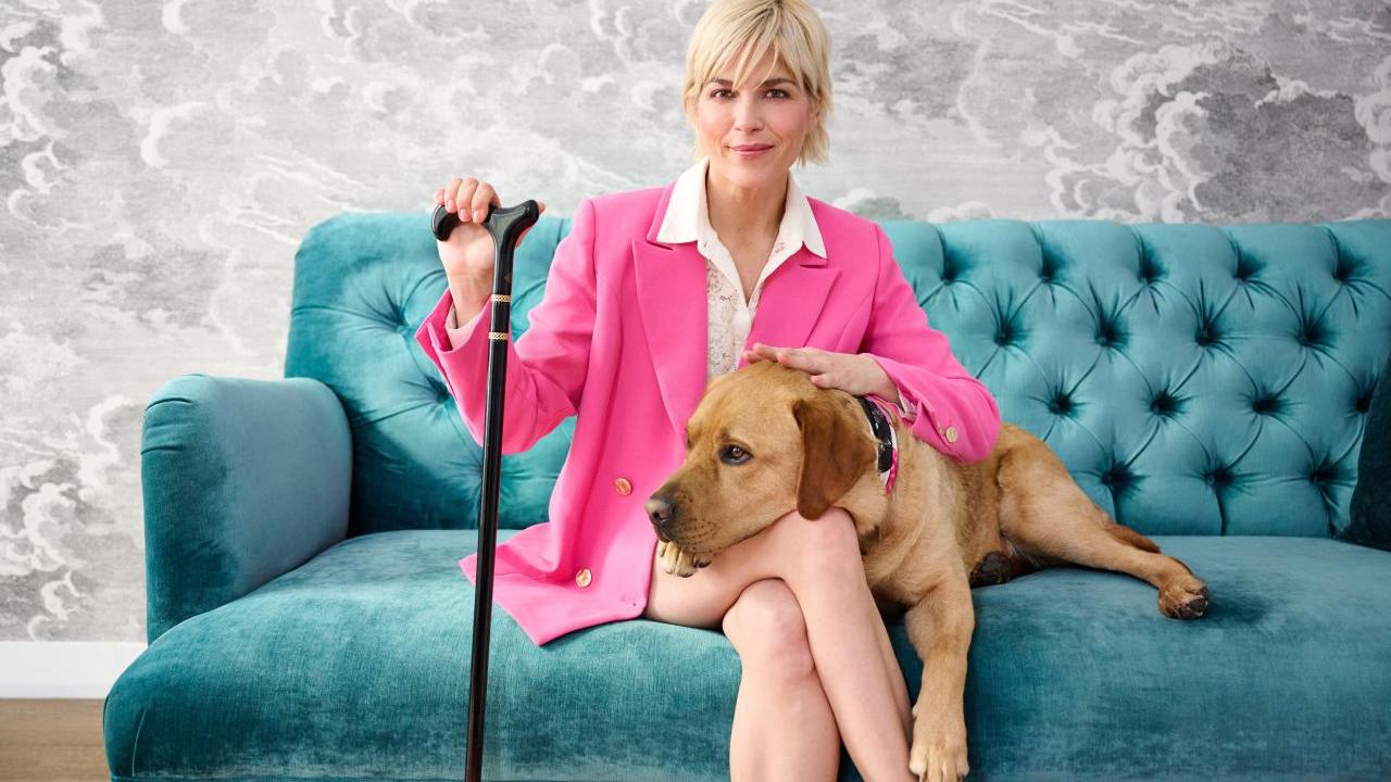 Selma Blair, QVC Brand Ambassador for Accessibility, sits on a plush teal sofa with her dog, Scout. She wears a hot pink suit and uses a black and gold cane.