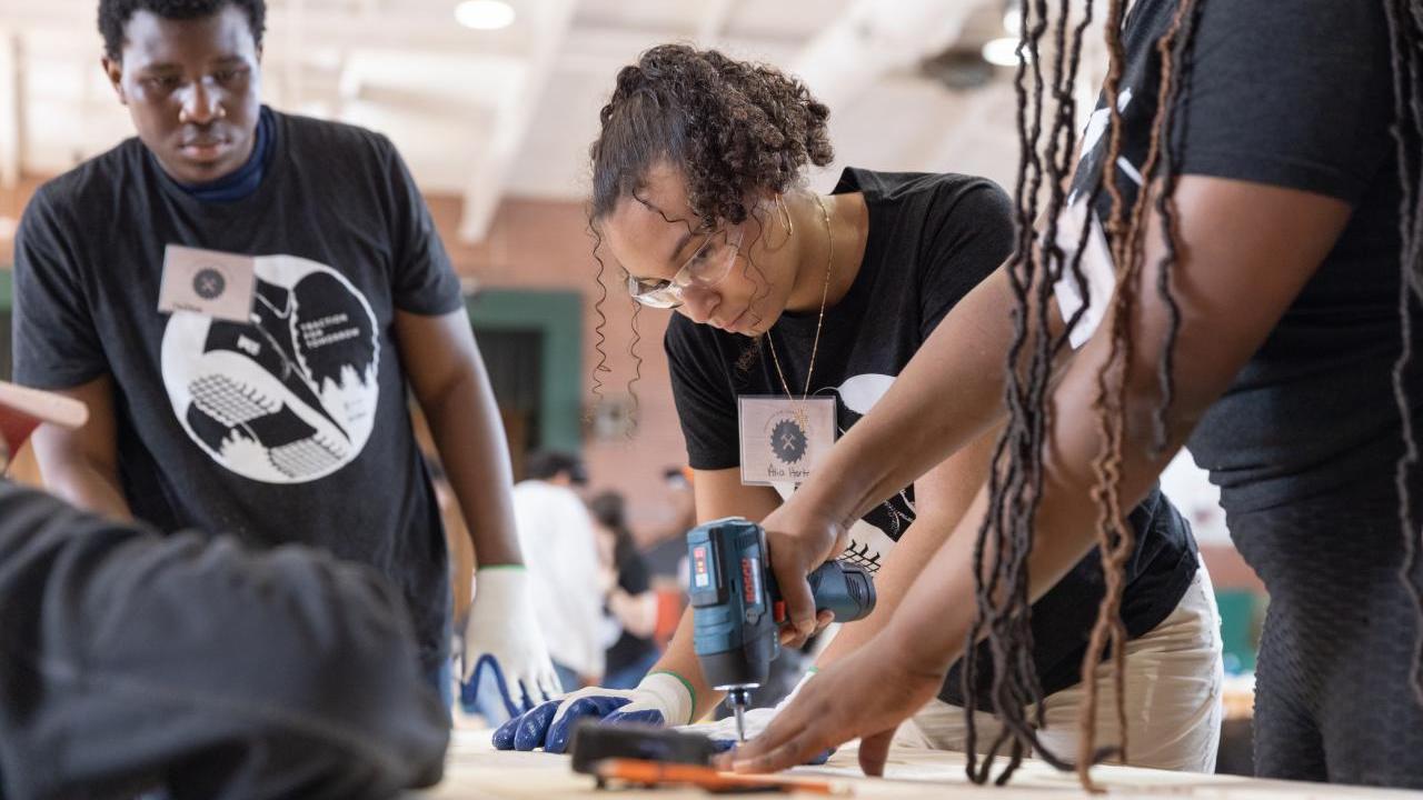 Person using a tool with another person helping and someone watching at the side