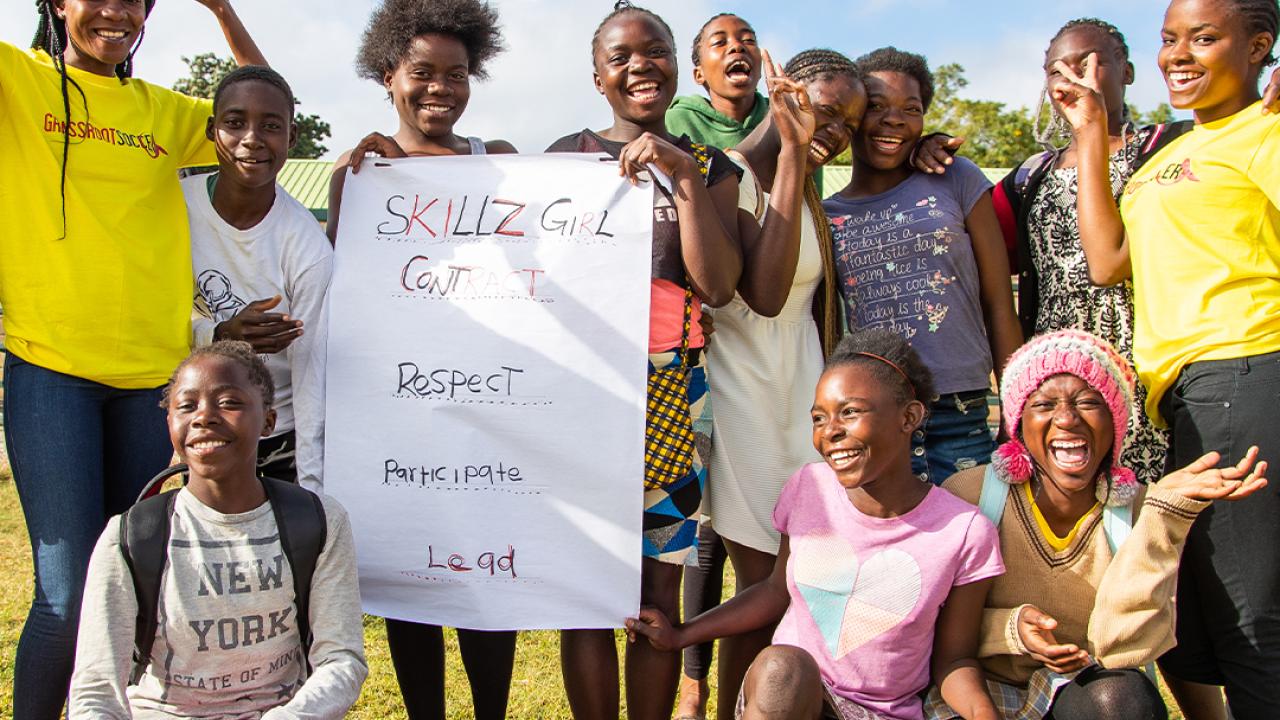 A group of youths smiling, posing for the camera on an open field, holding a sign "Skills Girl contract."