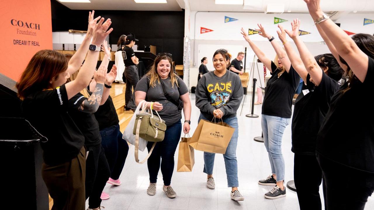 People standing in two lines with their arms raised while two women holding Coach shopping bags walk down the middle