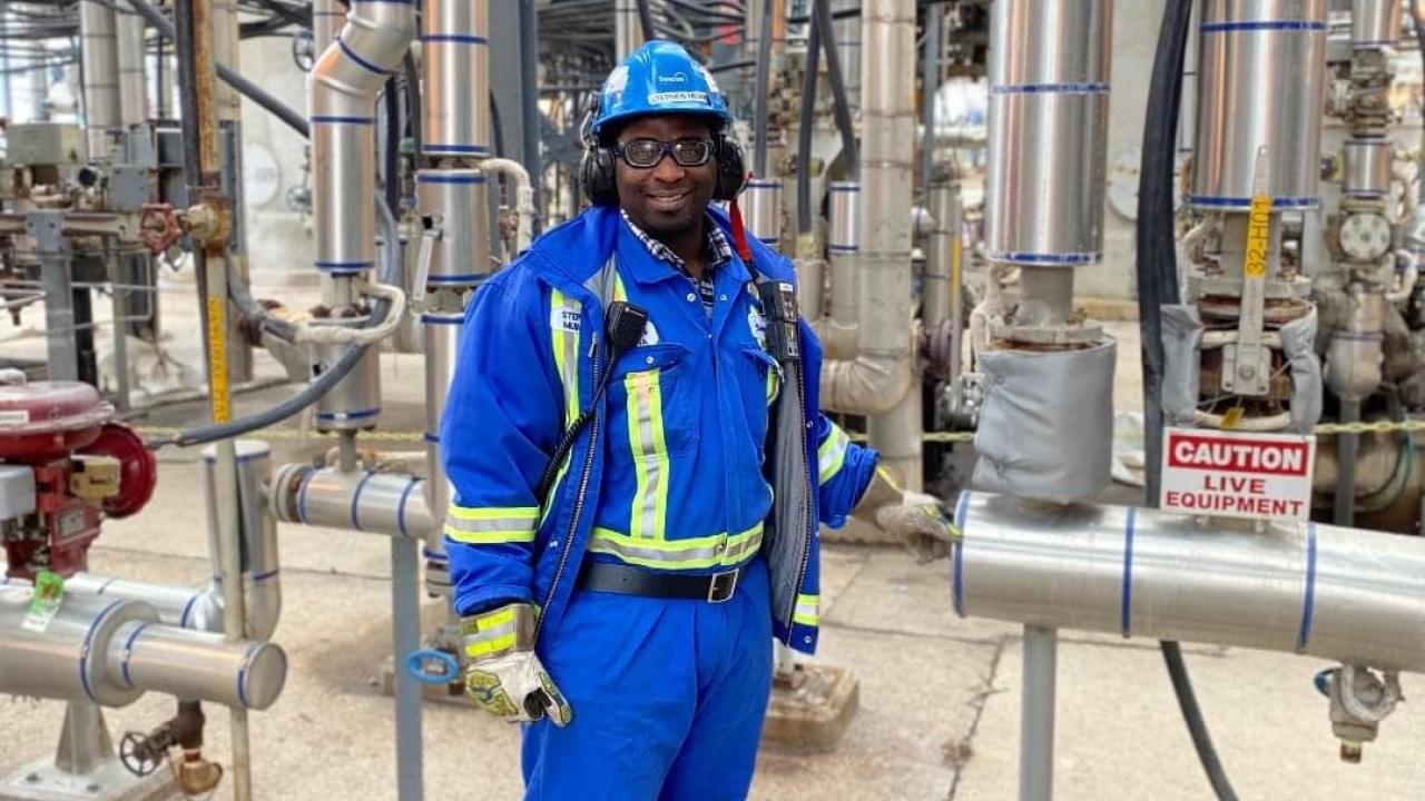 Stephen Muinda standing in an industrial complex, wearing protective clothing, hard hat, glasses, and ear muffs.