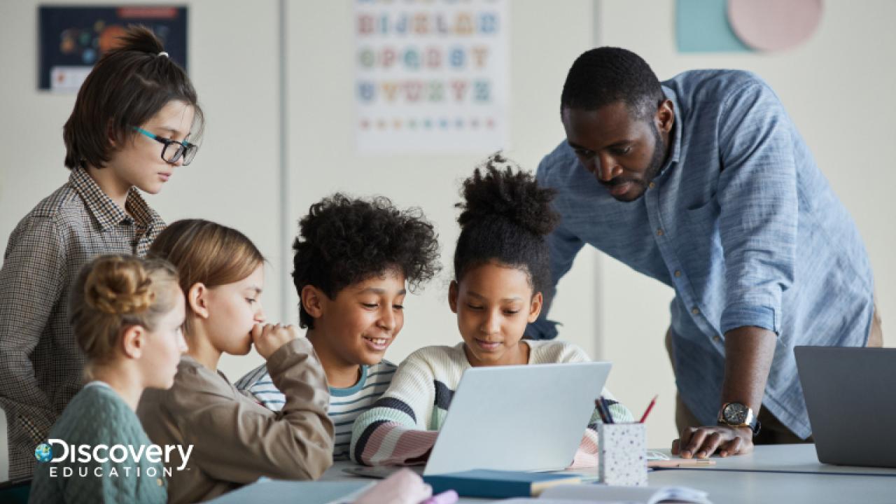 A teacher looking over students who are sat at a table working on a laptop