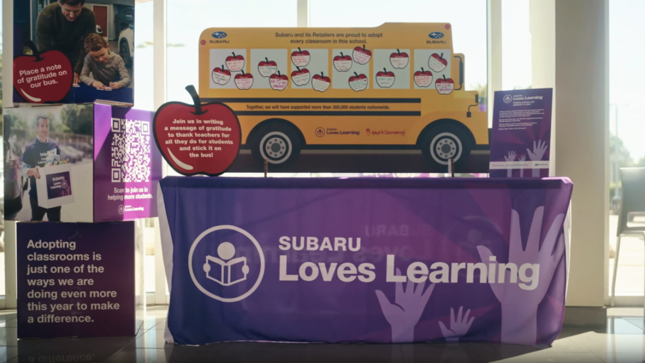 Still from a video showing a table with a paper school bus and signs about Subaru Loves Learning initiative