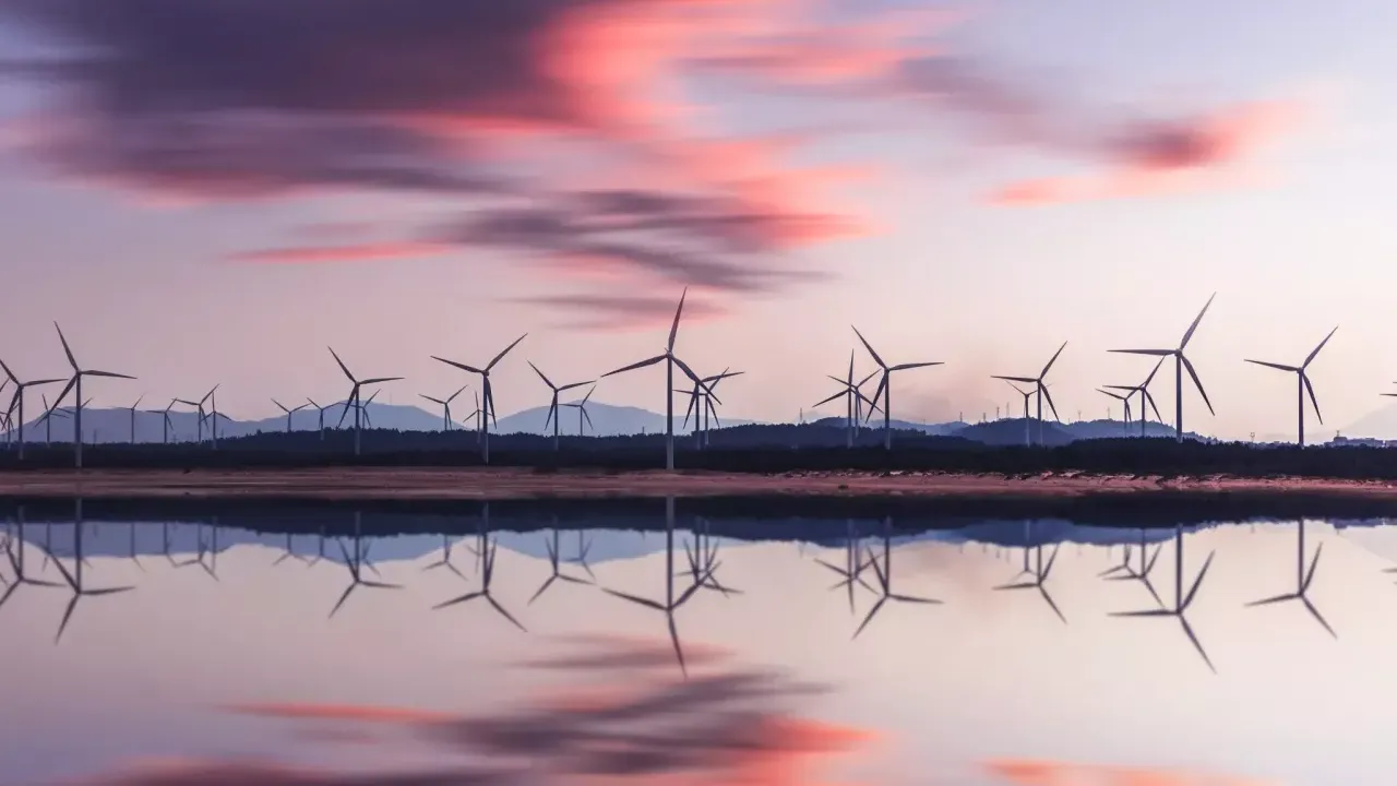 Rows of wind turbines in the distance, an open body of water in front reflecting clouds in a sunset.