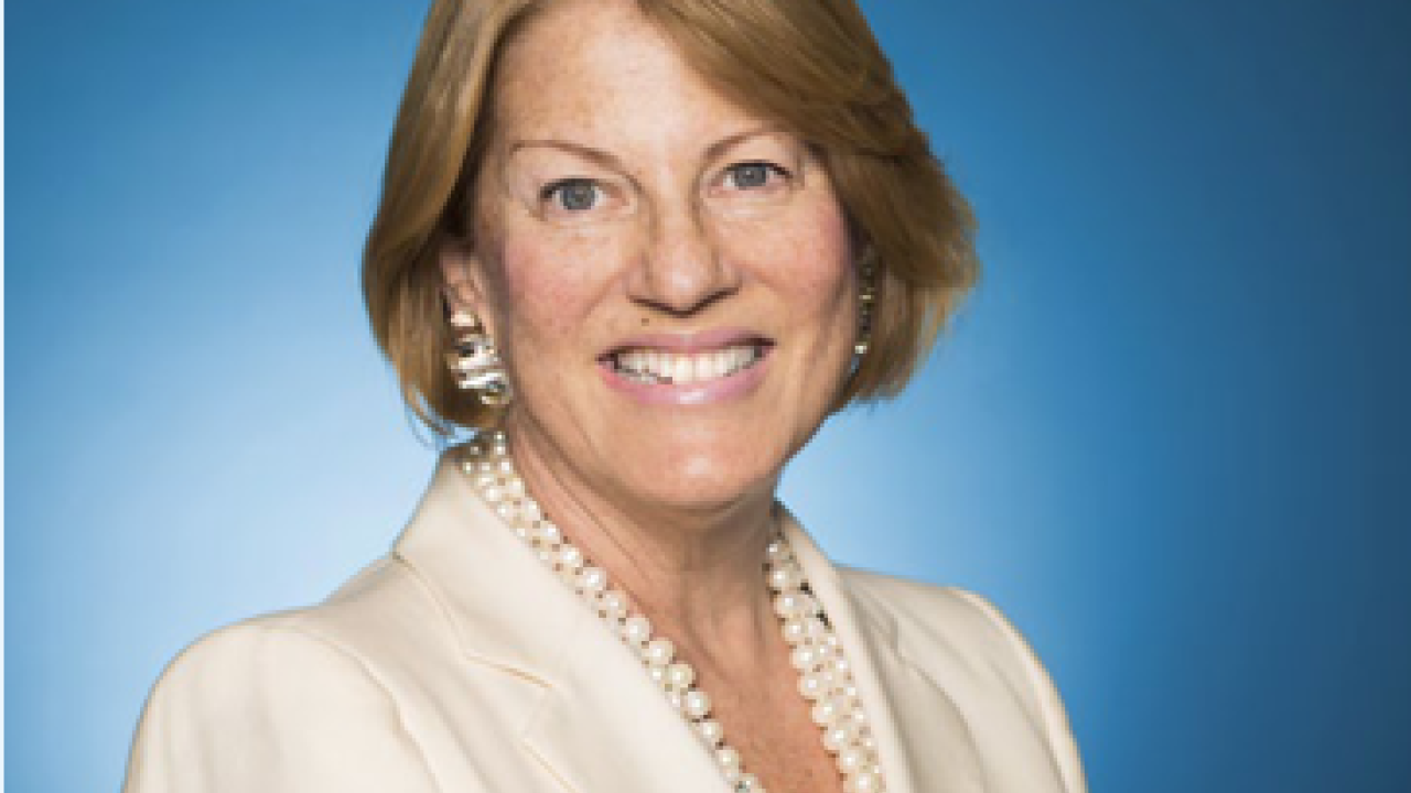 Profile of Sue Kronick. Blue background. Wearing a beige suit and pearl necklace