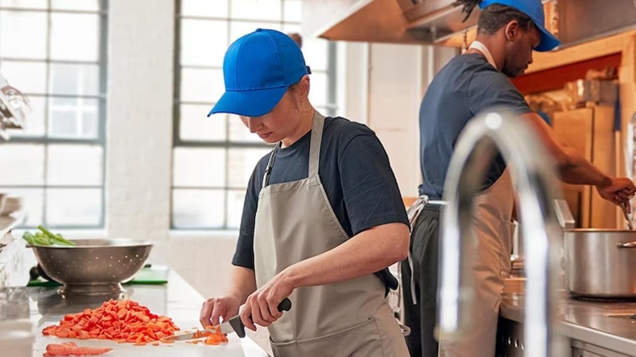 Two workers are shown preparing food in a restaurant. Both are wearing blue caps and aprons.