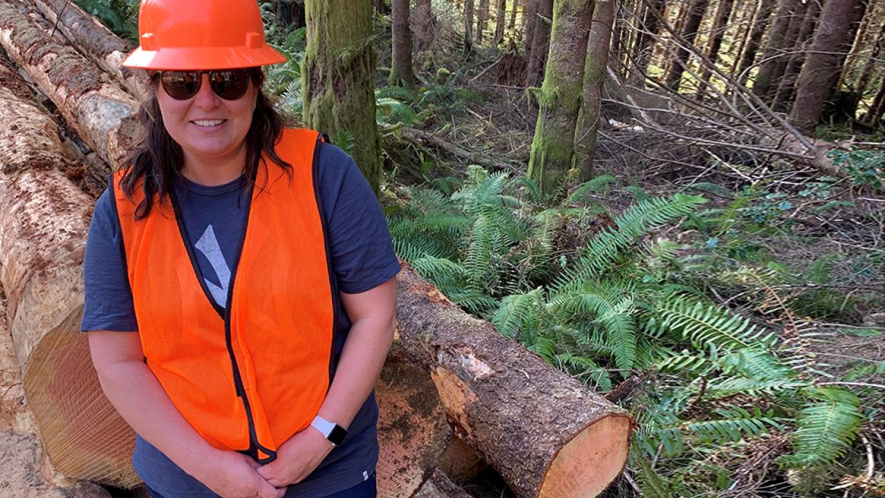 Tracie in front of cut timber in a forest wearing a safety hat and vest.