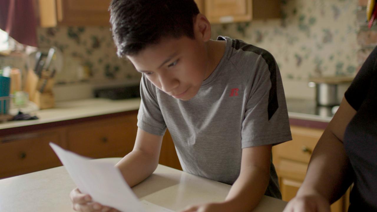 Young boy reading from a piece of paper at a kitchen table.