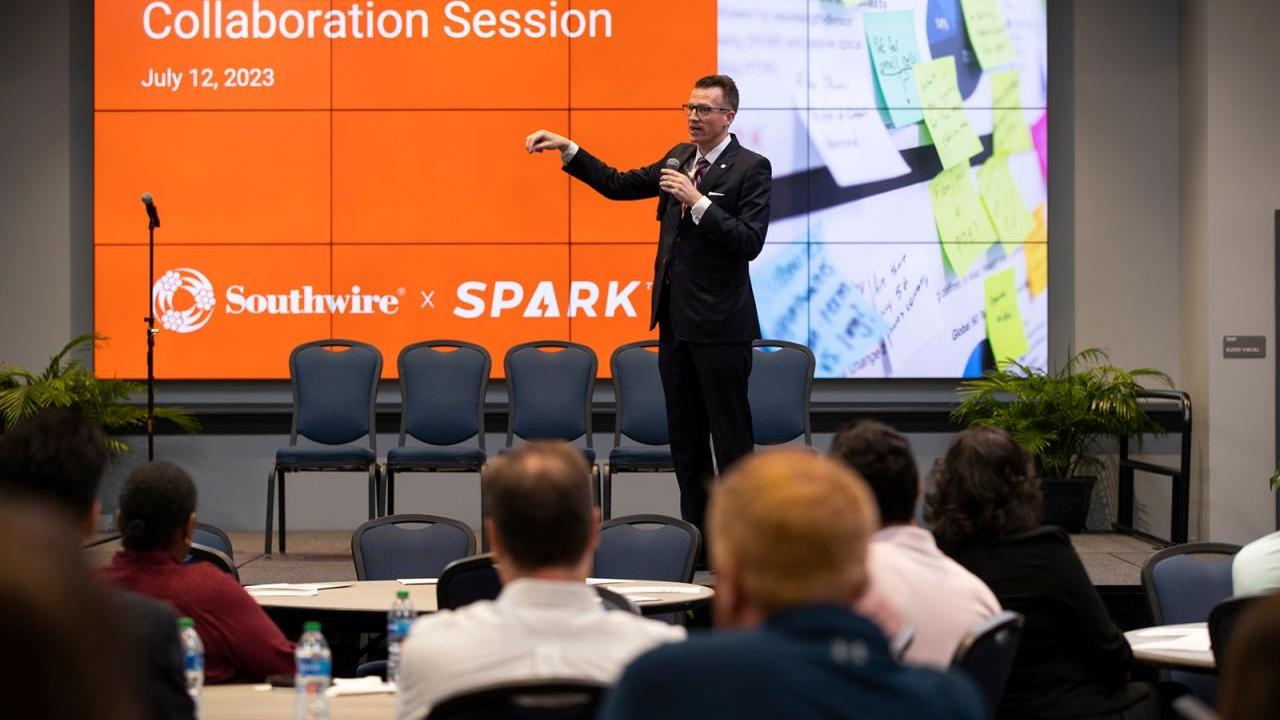 A speaker in front of a room of seated people. A digital screen behind with "UWG & Southwire Collaboration Session" 