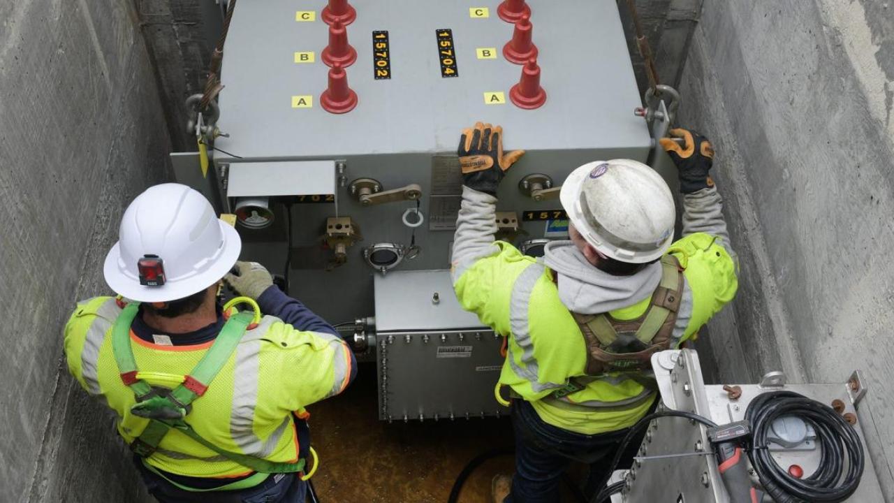 Two people in protective uniform working on a large electrical unit underground.