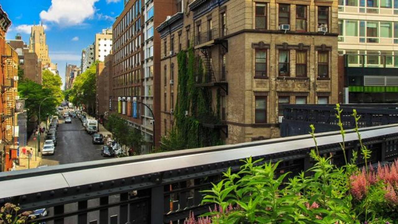 Panoramic view of an urban city with tall, close buildings. An elevated train line running through it. Greenery on the side.