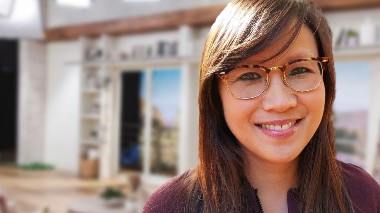 A photo of Virginia Nguyen with brown hair, wearing glasses and a maroon top while on-set at QVC Studios