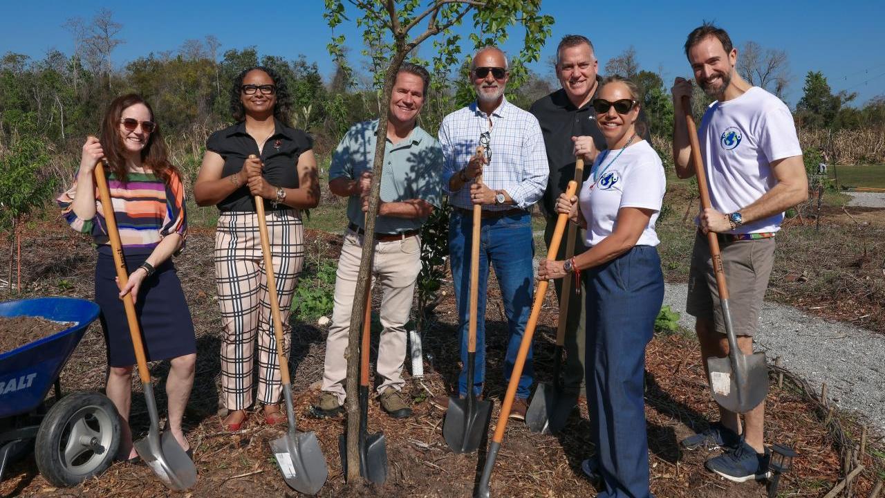 Seven people, including CEO of the Arbor Day Foundation, stand with shovels around newly planted tree. 